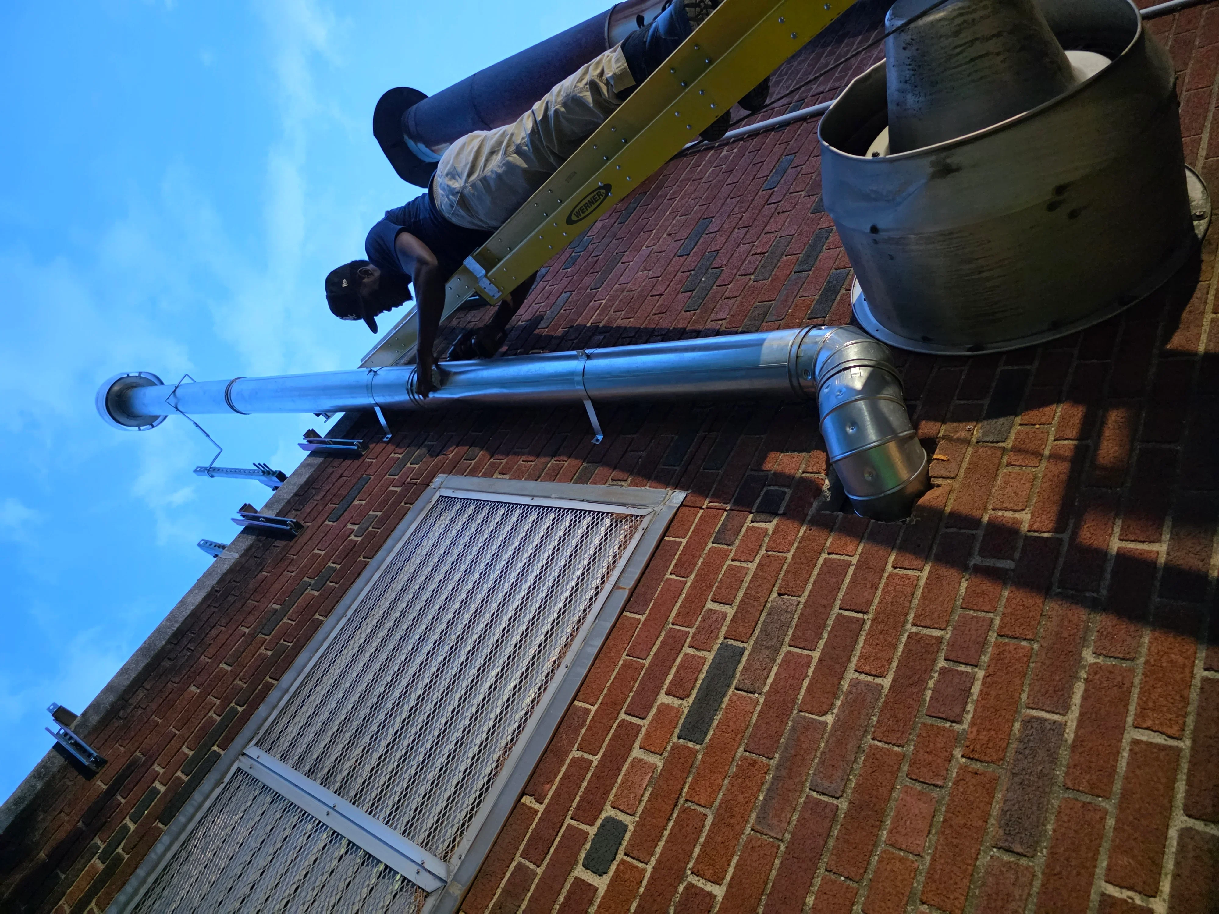 HVAC technician installing blue insulated ductwork and metal piping on brick building exterior